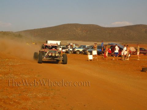 1990 Ford Bronco - long-travel off-road buggy races along a red dirt course past spectators in the desert.