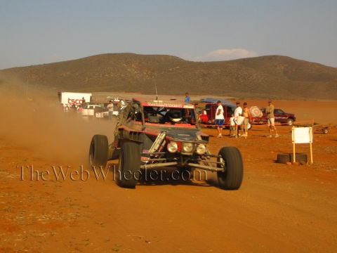 1990 Ford Bronco - black long-travel off-road buggy charges head-on down a dusty red dirt course past pit vehicles.