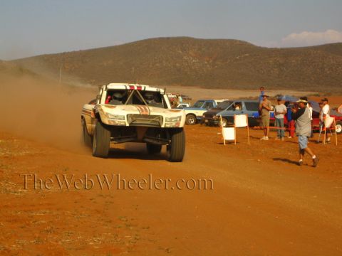 1990 Ford Bronco - white off-road race truck catches air on a red dirt course as spectators watch from the sidelines.
