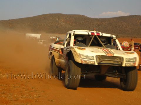 1990 Ford Bronco - white off-road race truck barrels head-on through red dust on a desert course.