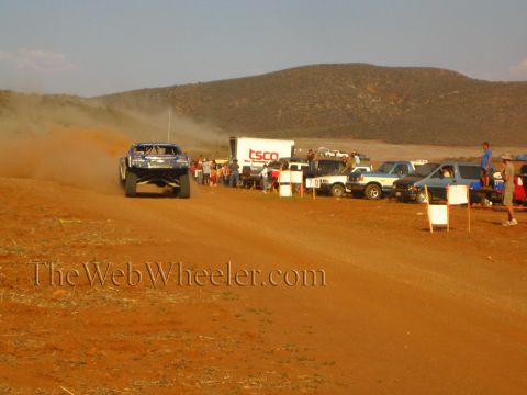 1990 Ford Bronco - blue off-road race truck trails dust on a red dirt course near a white TSCO box trailer.