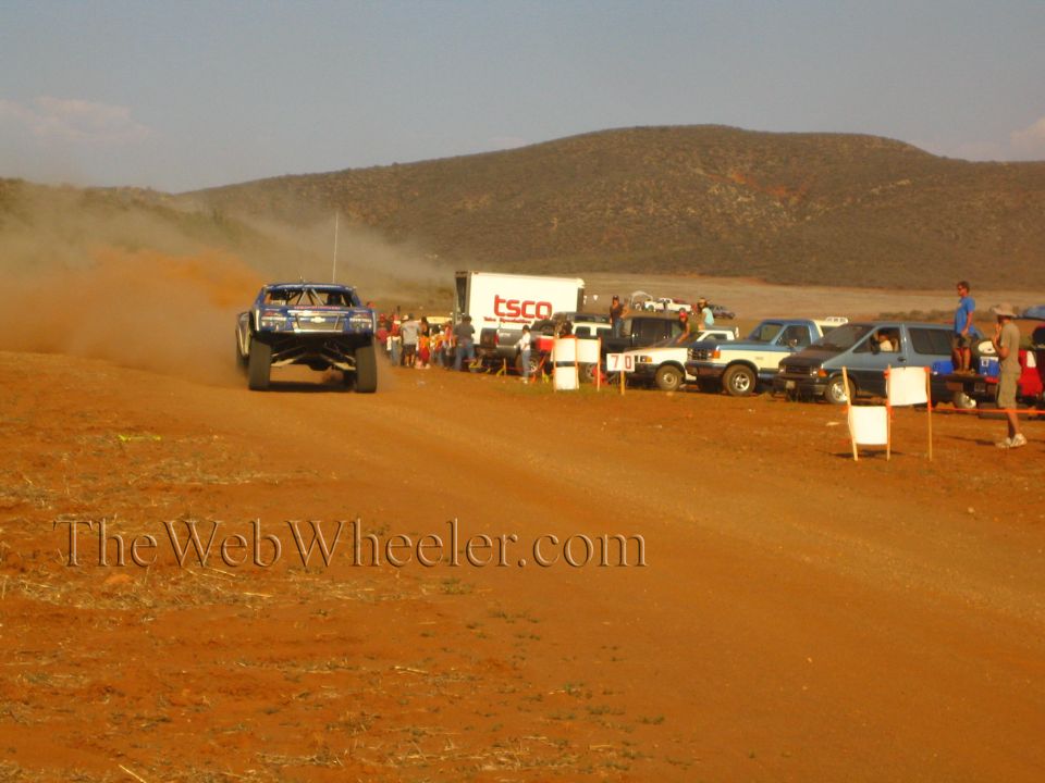 Blue off-road race truck on red dirt course past TSCO trailer