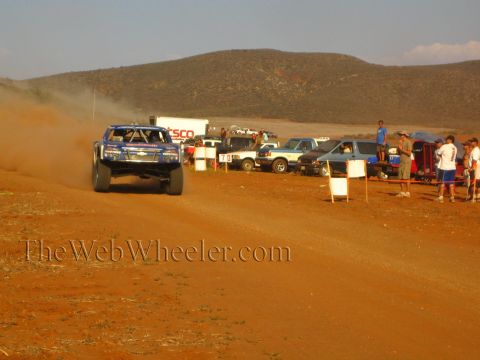 1990 Ford Bronco - blue off-road race truck kicks up dust on a red dirt course beside lined-up spectators and trucks.