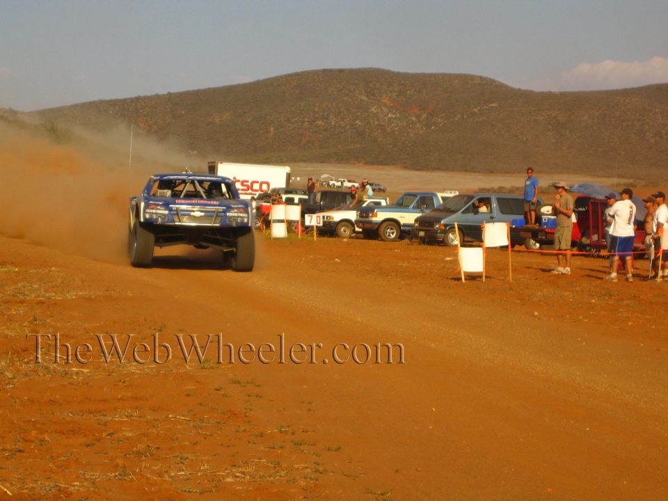 Blue off-road race truck racing on red dirt past spectators