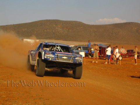 1990 Ford Bronco - blue off-road race truck with a Chevrolet front end charges head-on through red desert dust.