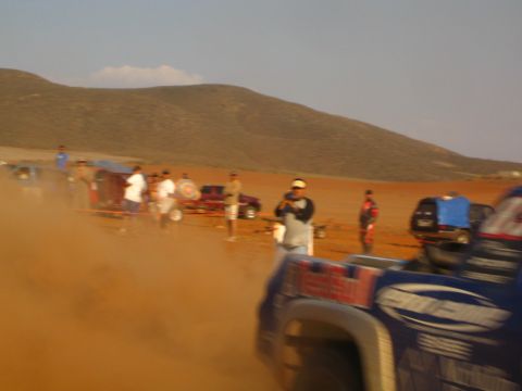 1990 Ford Bronco - From close behind, a blue off-road truck passes spectators in a blurry, dusty desert scene.