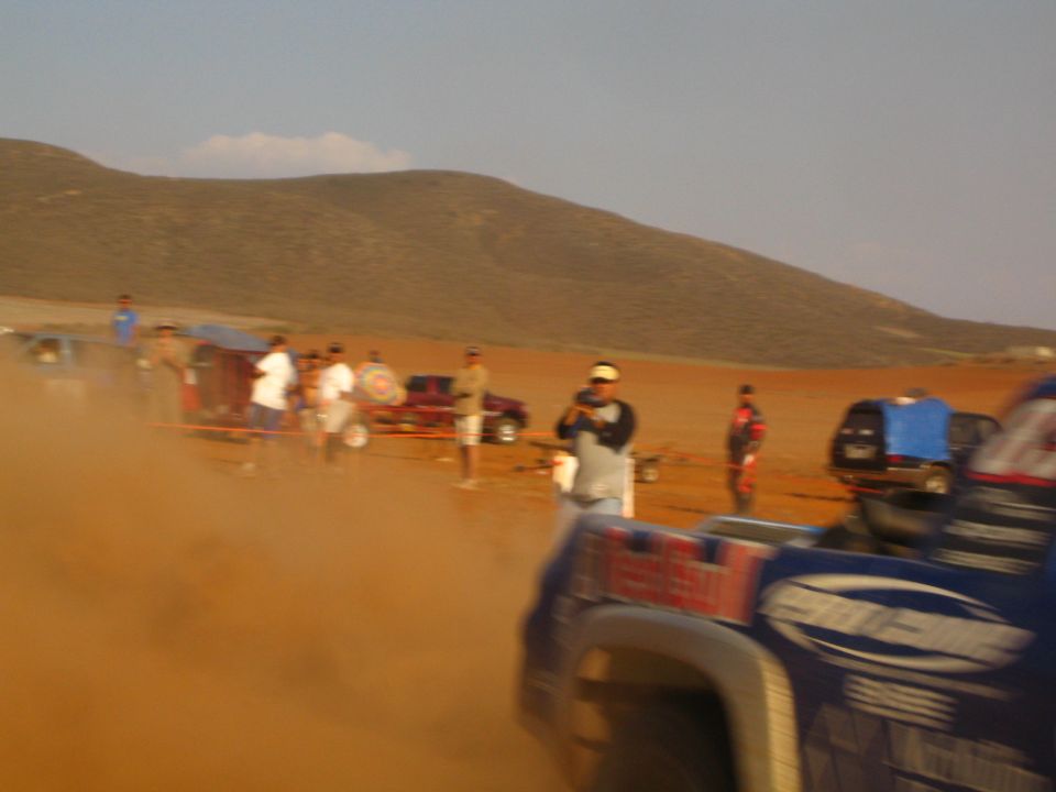 Blue off-road race truck passing spectators in blurry desert dust