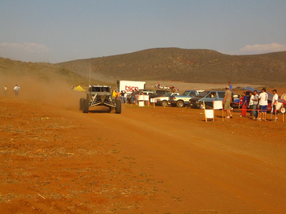Off-road buggy on red dirt course past TSCO trailer and crowd