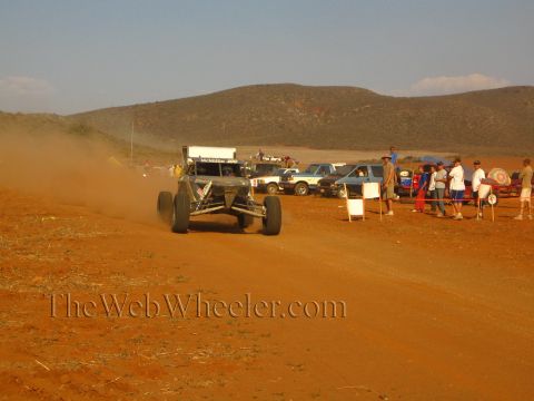 1990 Ford Bronco - white-roofed off-road buggy trails dust on a red dirt course as spectators watch nearby.