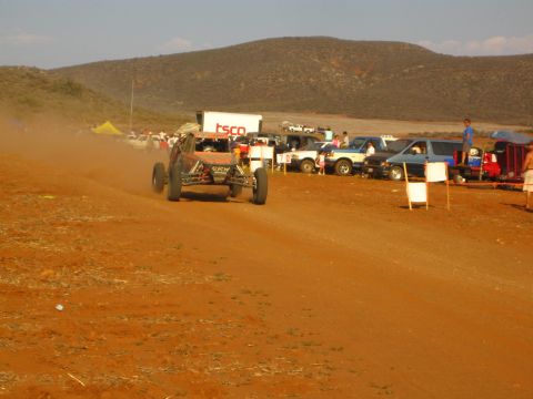 1990 Ford Bronco - off-road buggy trails dust on a red dirt course past parked trucks and a TSCO trailer.
