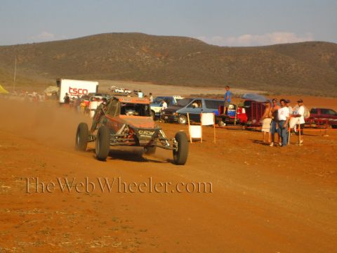 1990 Ford Bronco - dark off-road buggy kicks up dust on a red dirt course as spectators watch beside parked vehicles.