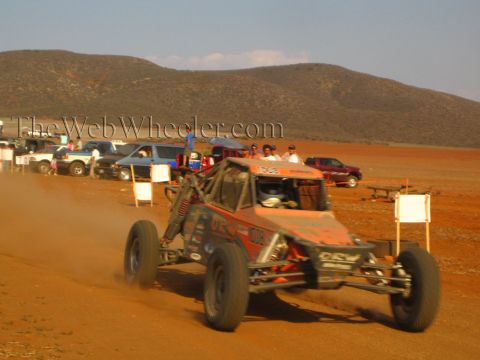 1990 Ford Bronco - red off-road buggy carves through red dust on a desert course past a row of parked vehicles.