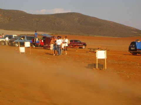 1990 Ford Bronco - Spectators and parked trucks gather in a dusty desert pit area dotted with white course-marker boards.
