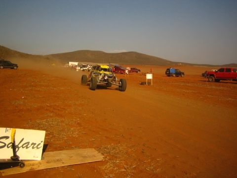 1990 Ford Bronco - yellow off-road buggy speeds along a red dirt course beyond a Safari course sign and parked trucks.