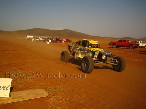 1990 Ford Bronco - blue and yellow tube-frame off-road race buggy kicks up dust on a red dirt course as spectator trucks line the background.