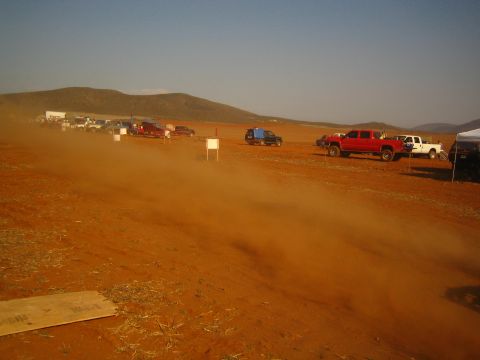 1990 Ford Bronco - dust cloud drifts across a dirt staging area where red and white pickup trucks and other vehicles park near desert hills.
