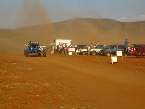 1990 Ford Bronco - blue race buggy speeds past a line of spectators, trucks, and a box truck along a dusty red dirt course.