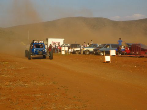 1990 Ford Bronco - blue tube-frame off-road buggy rounds a dirt course past parked pickups and a small crowd of spectators.