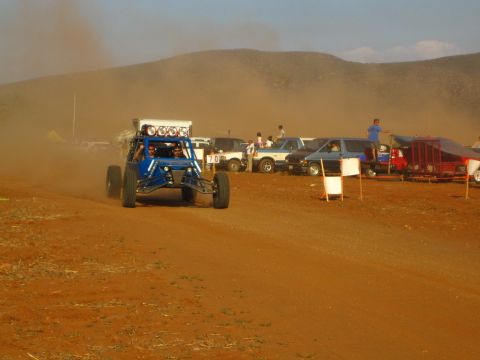 1990 Ford Bronco - blue off-road race buggy fitted with a roof light bar drives a red dirt course near desert hills and spectators.