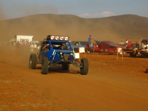 1990 Ford Bronco - blue off-road race buggy with a roof light bar races a dusty dirt course past a box truck and spectators.