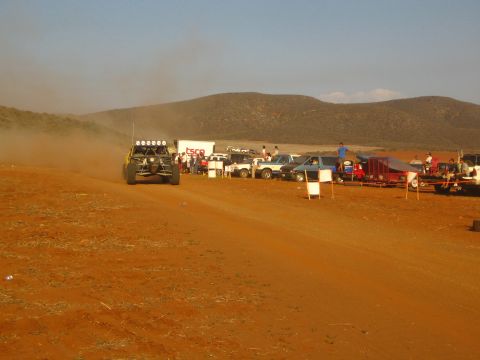 1990 Ford Bronco - dark off-road buggy with a roof light bar heads down a red dirt course toward a pit area lined with trucks.