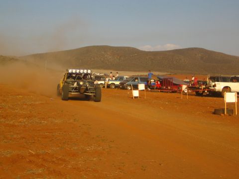 1990 Ford Bronco - off-road race buggy with a roof light bar drives away on a red dirt course past parked vehicles and spectators.