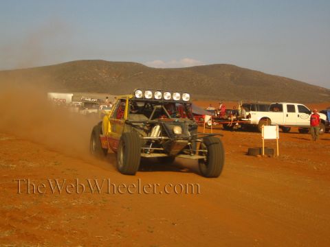 1990 Ford Bronco - yellow tube-frame off-road buggy with a roof light bar races a red dirt course as a white pickup waits in the pit.