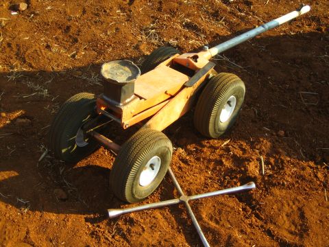 1990 Ford Bronco - homemade wooden four-wheel cart or jack dolly rests on red dirt beside a chrome cross-style lug wrench.