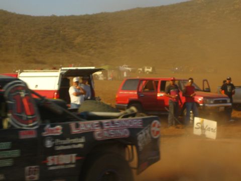 1990 Ford Bronco - black race vehicle marked Lake Elsinore Casino sits in a dusty pit area as spectators and red trucks gather behind.