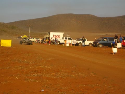 1990 Ford Bronco - TSCO box truck, pickups, and a yellow course flag mark a dusty red dirt staging area below desert hills.