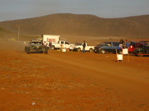 1990 Ford Bronco - off-road race buggy speeds along a red dirt course past parked trucks and a TSCO box truck in the pits.