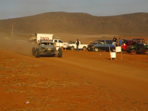 1990 Ford Bronco - tube-frame off-road buggy races a red dirt course past parked pickups, a box truck, and a green UTV.