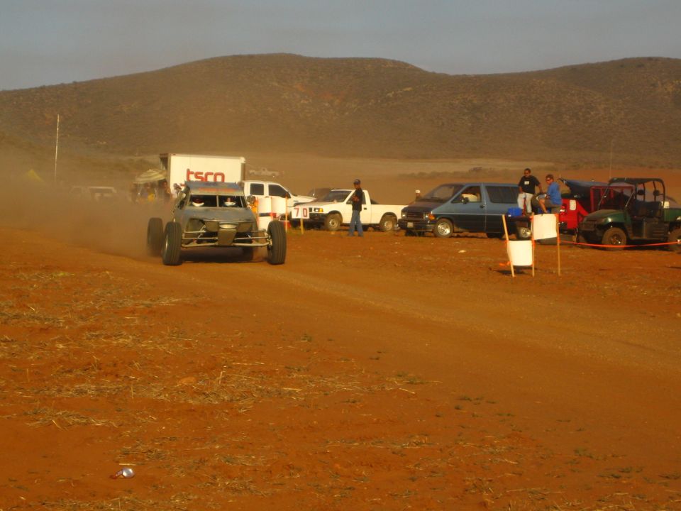 Tube-frame buggy on red dirt course past trucks and a UTV