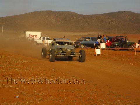 1990 Ford Bronco - bare-metal tube-frame off-road buggy kicks up dust on a red dirt course beside a pit line of trucks and a UTV.