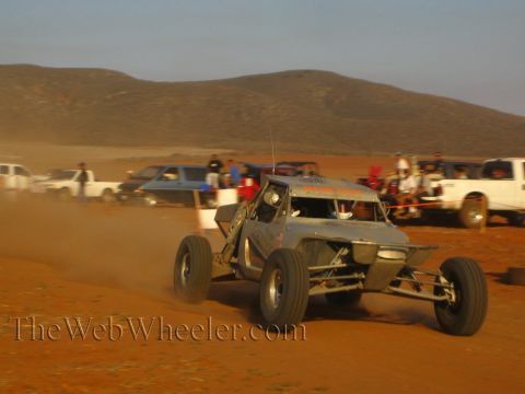 1990 Ford Bronco - gray tube-frame off-road race buggy drives fast across a red dirt course as spectators and trucks watch behind.