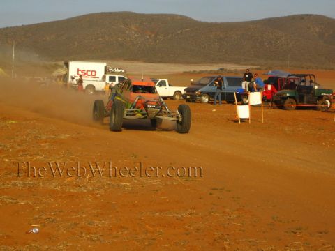 1990 Ford Bronco - orange and green tube-frame off-road buggy races a dusty red dirt course past pit trucks and a UTV.