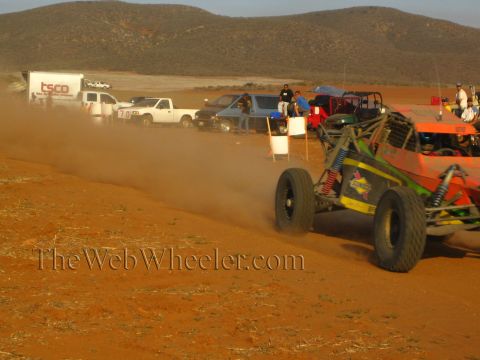 1990 Ford Bronco - orange tube-frame off-road buggy with tall coilover shocks corners on red dirt near a pit line of trucks.