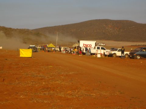 1990 Ford Bronco - long pit line of trucks, a TSCO box truck, and a yellow flag stretches across a dusty red dirt desert area.