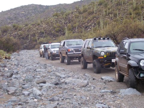 1991 Ford Bronco - Multiple off-road SUVs navigate a rough, rocky trail in a desert landscape. Supermotors.net logo is present.