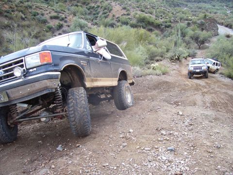 1991 Ford Bronco - A lifted Ford Bronco navigates a dusty, off-road trail with other vehicles in the background.