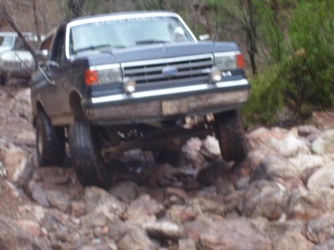 1991 Ford Bronco - A dark blue Ford Bronco navigates a rocky trail, with another vehicle partially visible in the background.
