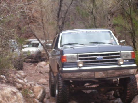 1991 Ford Bronco - A dark blue 1991 Ford Bronco navigates a rough, rocky terrain, with another vehicle visible in the background.