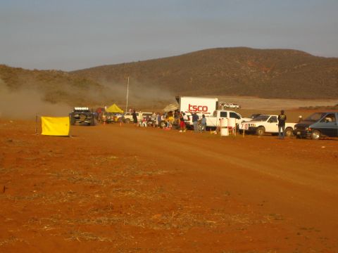 1990 Ford Bronco - Spectators and a row of pickups gather around a TSCO box truck in a dusty red dirt desert race pit.