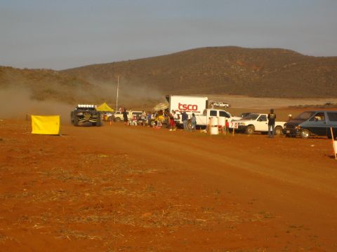 1990 Ford Bronco - off-road buggy sits at the edge of a busy pit row with a TSCO box truck and pickups on red desert dirt.