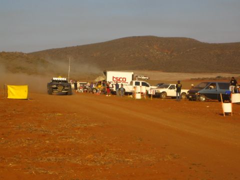 1990 Ford Bronco - off-road buggy with roof lights waits beside a pit crowd, a TSCO box truck, and pickups on red desert dirt.