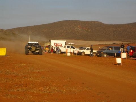 1990 Ford Bronco - off-road buggy with a roof light bar idles near a TSCO box truck and pit pickups on a red dirt desert course.