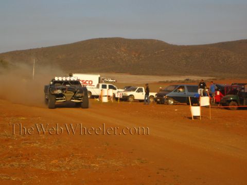 1990 Ford Bronco - off-road desert race buggy kicks up dust on a red dirt course while a TSCO box truck and pit vehicles line the route at dusk.