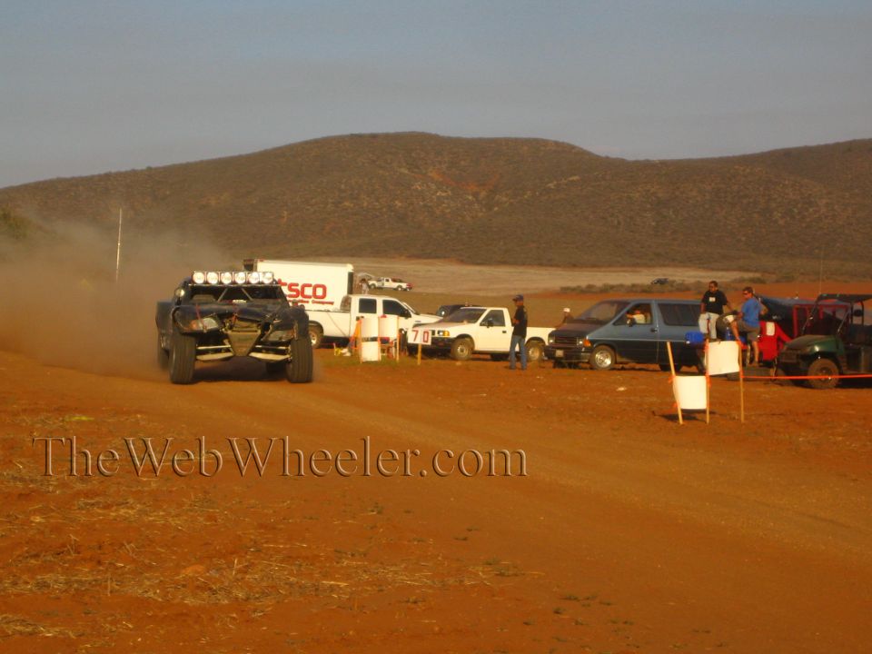 Off-road race buggy speeding on red dirt course past trucks and spectators at dusk