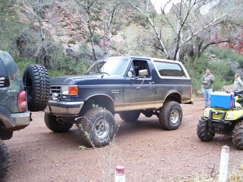 1991 Ford Bronco - A lifted 1991 Ford Bronco is parked off-road with other vehicles and people nearby.
