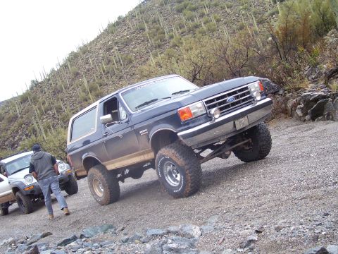1991 Ford Bronco - A dark blue Ford Bronco with a lifted suspension navigates a rocky, off-road trail in a desert landscape.